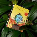 A book titled Liebe Erde by War and Peas rests among the green leaves of a plant. The cover illustration features a cartoon Earth wearing a small hat, sweating nervously while holding a burning letter, surrounded by flames. A red circular sticker on the cover indicates the inclusion of new and exclusive comic strips. The book is published by Edition Moderne. The vibrant yellow background of the cover contrasts with the green foliage surrounding it.