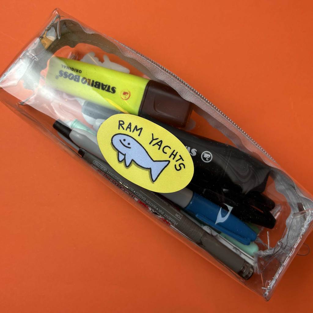 A transparent pencil case filled with pens, markers, and highlighters. A yellow “RAM YACHTS” sticker with a cartoon fish is stuck on the outside of the case.