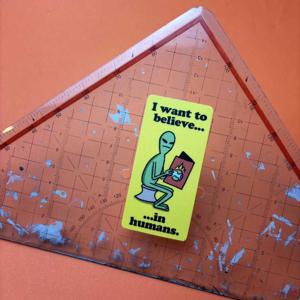 A bright yellow sticker with a humorous alien graphic and the text 'I want to believe... in humans' placed on a transparent ruler, ideal for those who love witty and eye-catching stickers.