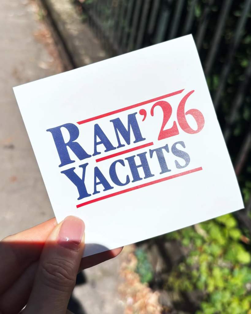 A hand holds up a square white "RAM YACHTS '26 " sticker outdoors on a sunny sidewalk, a black metal fence and green foliage blurred in the background. The design mimics a US political campaign logo.