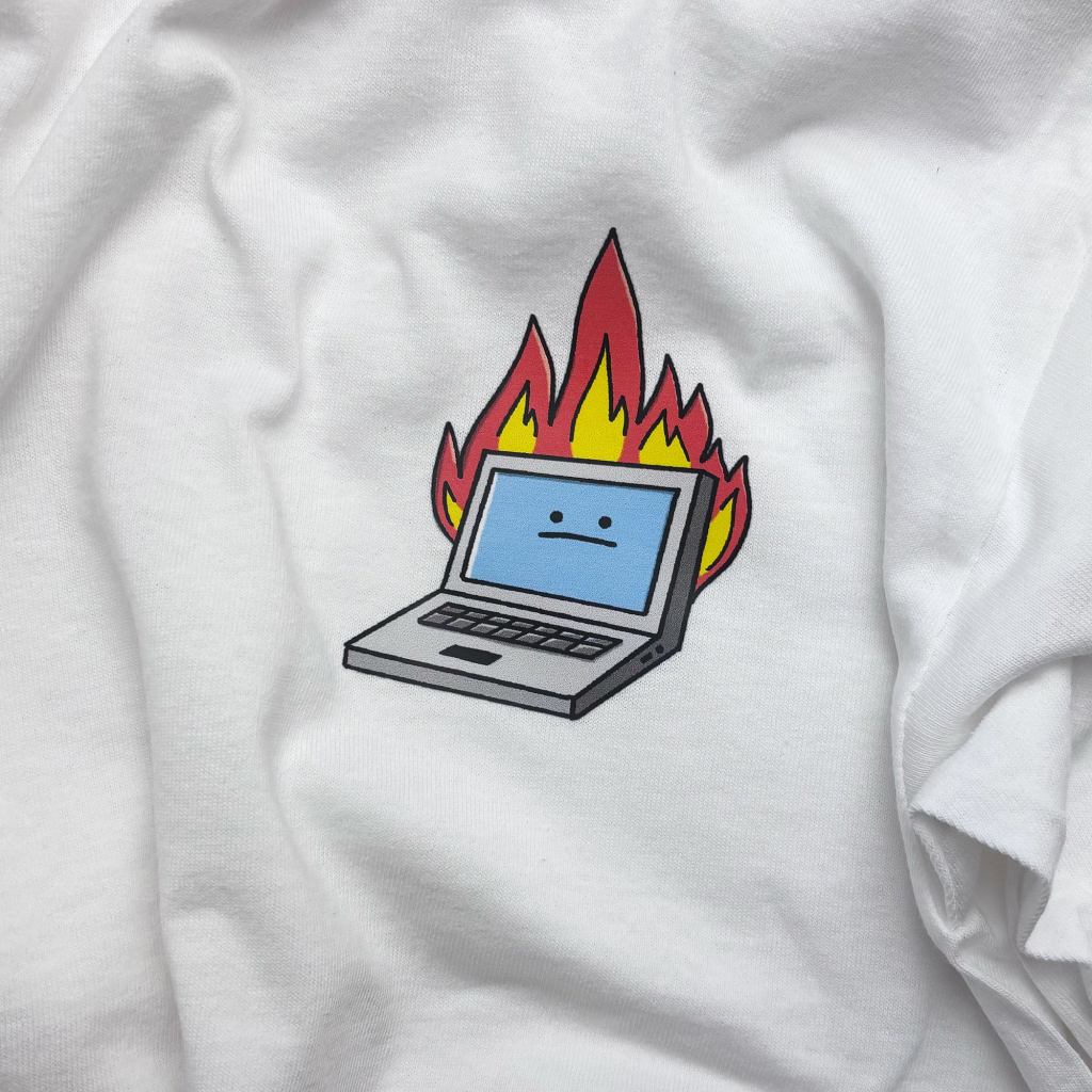 A white t-shirt with a War and Peas textile sticker of a cartoon laptop engulfed in red and yellow flames. The laptop's blue screen wears a flat, unimpressed expression. Applied to soft cotton fabric, this "Laptop on Fire" iron-on textile sticker is a humorous nod to burnout and digital overwhelm.