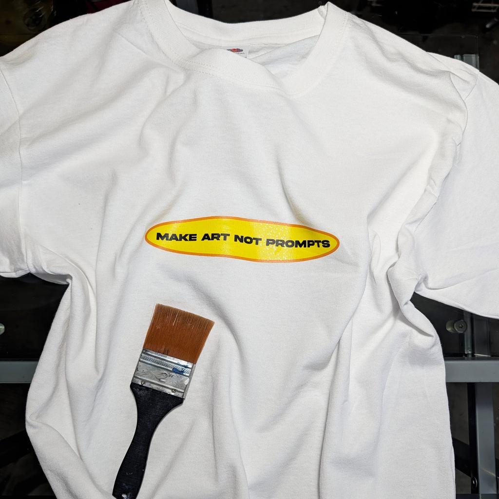 A white cotton t-shirt with a yellow oval textile sticker reading "MAKE ART NOT PROMPTS" in bold black capitals, a well-used 2-inch paintbrush resting on the fabric beside it. This War and Peas "Make Art Not Prompts" iron-on sticker is a pro-artist, anti-AI slogan celebrating handmade creativity.