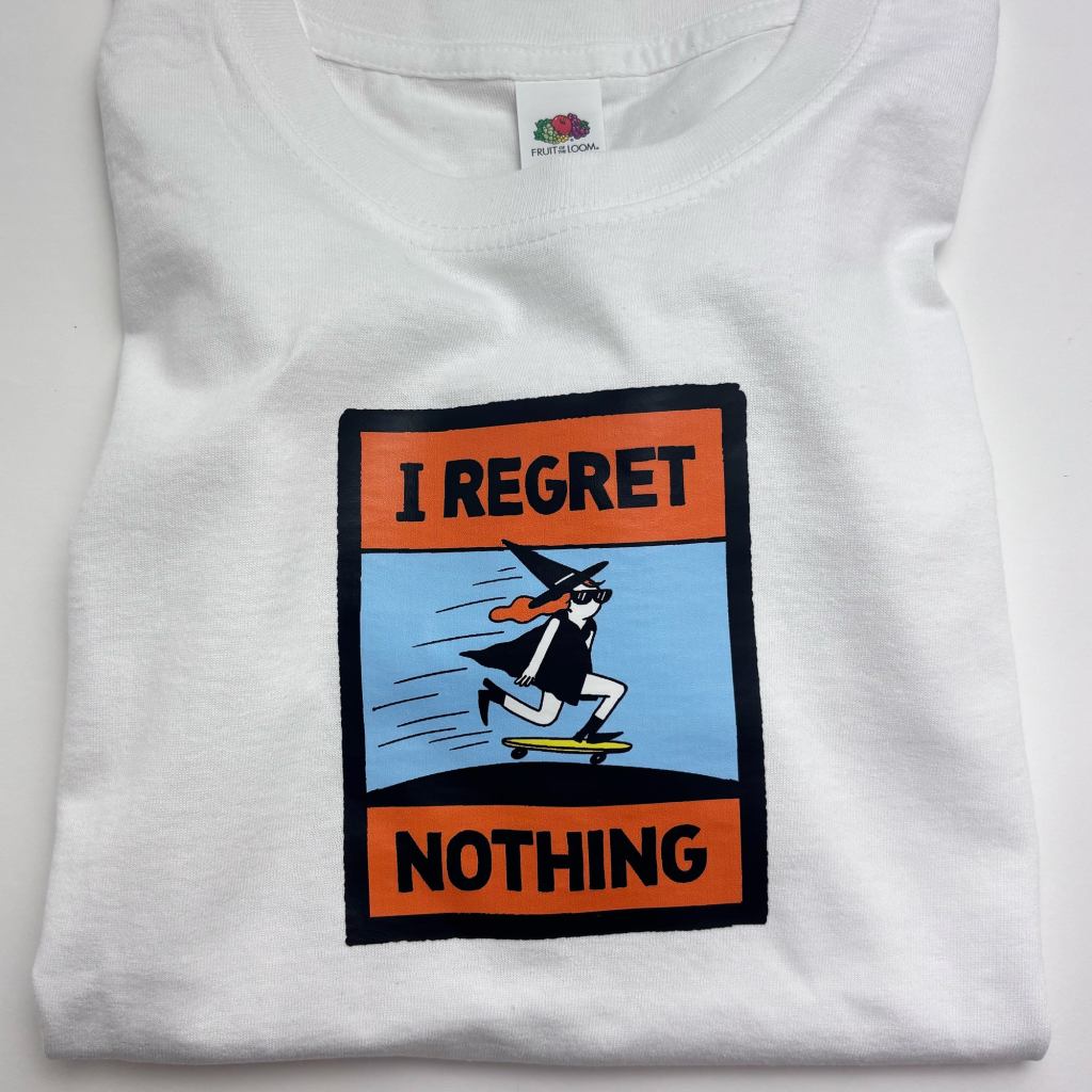 A folded white t-shirt with a comic-style textile sticker reading "I REGRET NOTHING" in bold black letters on orange panels. In the blue center panel, a witch in a pointed hat and sunglasses rides a yellow skateboard downhill, cape flying. This War and Peas "I Regret Nothing" iron-on sticker is a playful, carefree statement design.